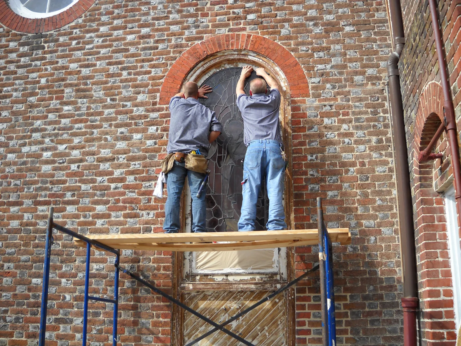 Workmen on scaffolding fitting a stained glass window into the church's brick wall